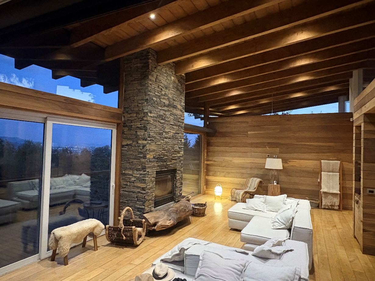 Casa Araucanía — Living con chimenea de piedra, vigas de madera a la vista y ventanales